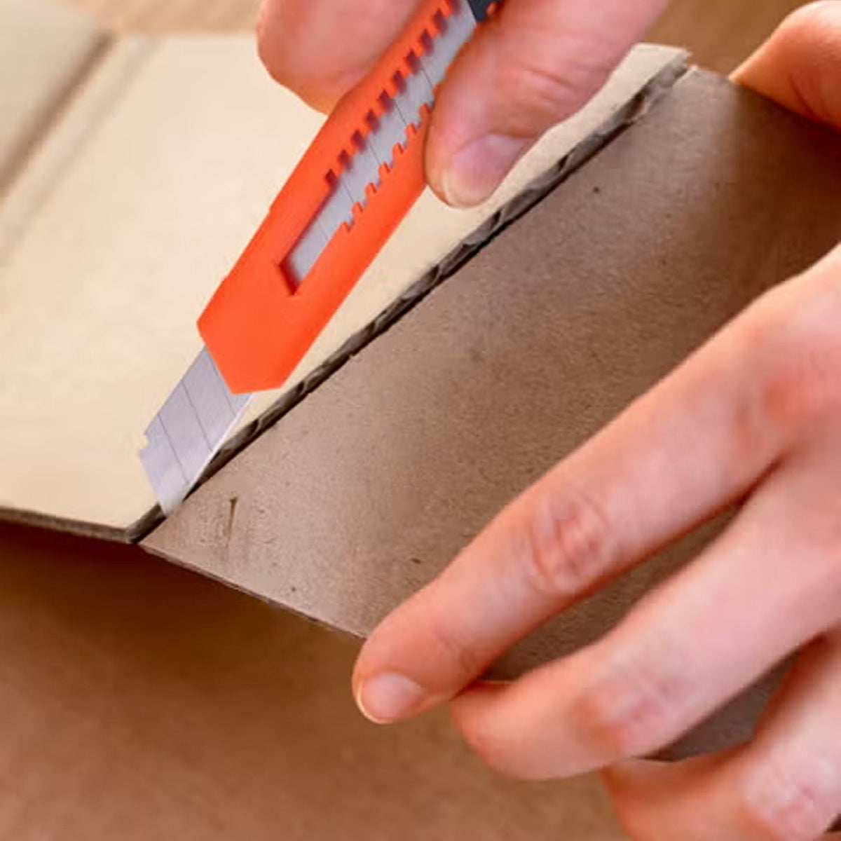 A hand uses an orange utility knife to cut a piece of cardboard, showing the blade in action.
