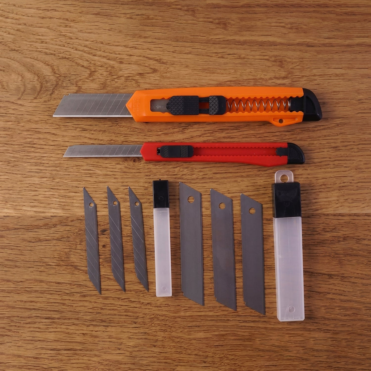 Two utility knives, one orange and one red, with extra blades and storage cases, arranged on a wooden table.