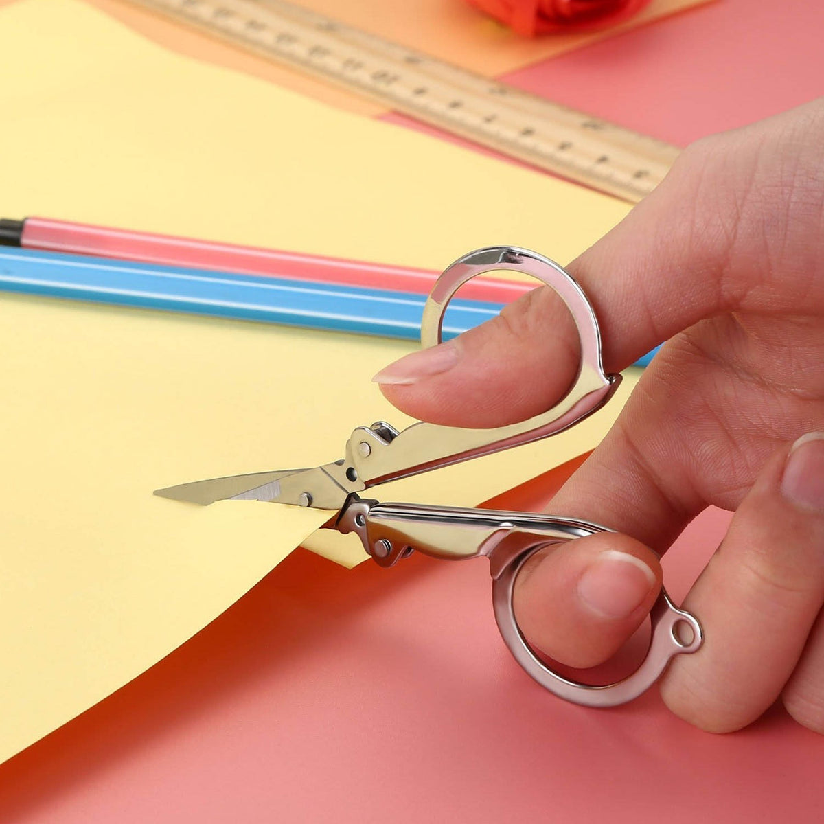 A hand uses open silver folding scissors to cut yellow paper, with a ruler and pencils in the background.