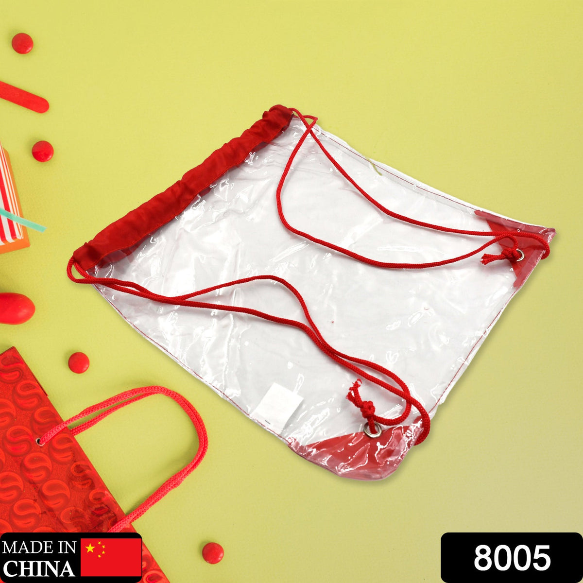 Clear drawstring bag with red cords and accents, lying flat on a bright yellow surface with red decorative items.