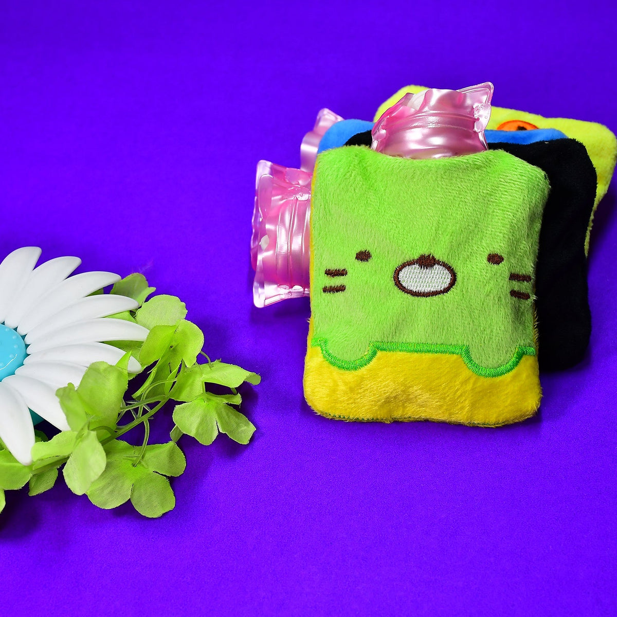 Two green and yellow kitty hot water bags with pink tops are on a purple background with a white flower.