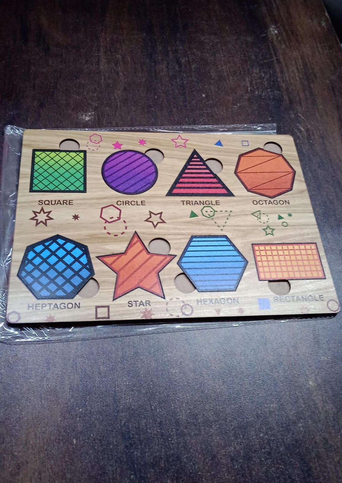 The wooden shape sorting puzzle with all colorful pieces in place is shown in plastic packaging on a dark surface.