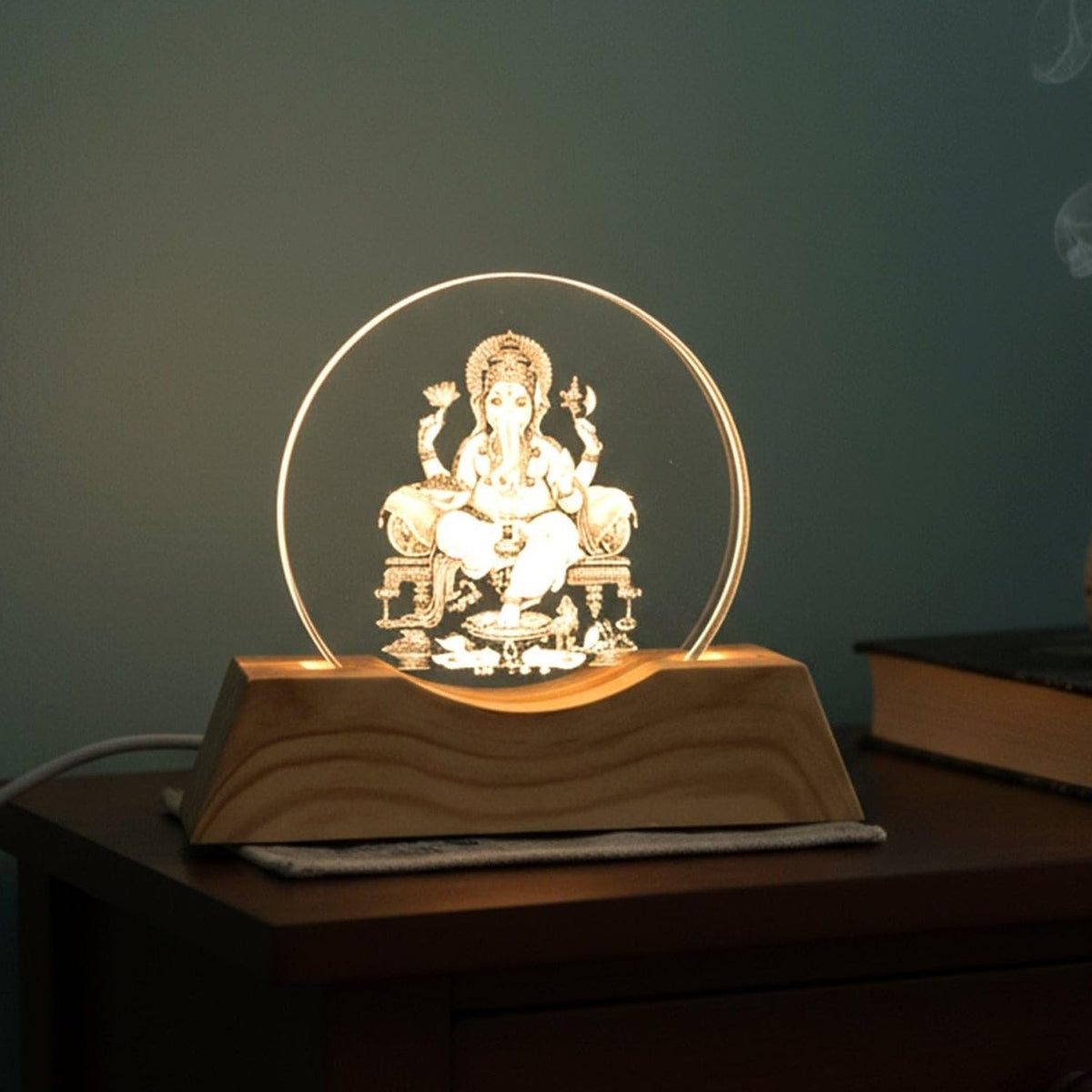 The Ganesha 3D illusion lamp glows warmly on a dark wooden nightstand in a cozy room.