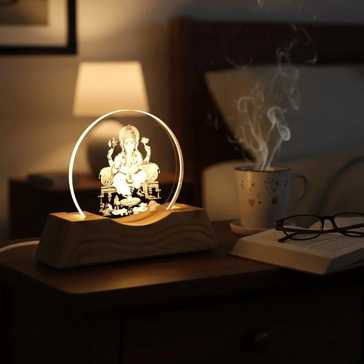 Ganesha 3D illusion lamp on a bedside table with a steaming mug, book, and glasses nearby.