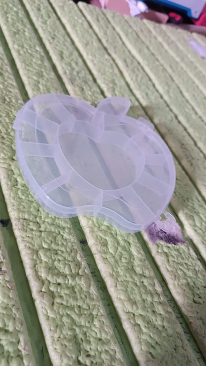A transparent apple-shaped organizer box with a heart-shaped center is placed on a textured green surface, viewed from a