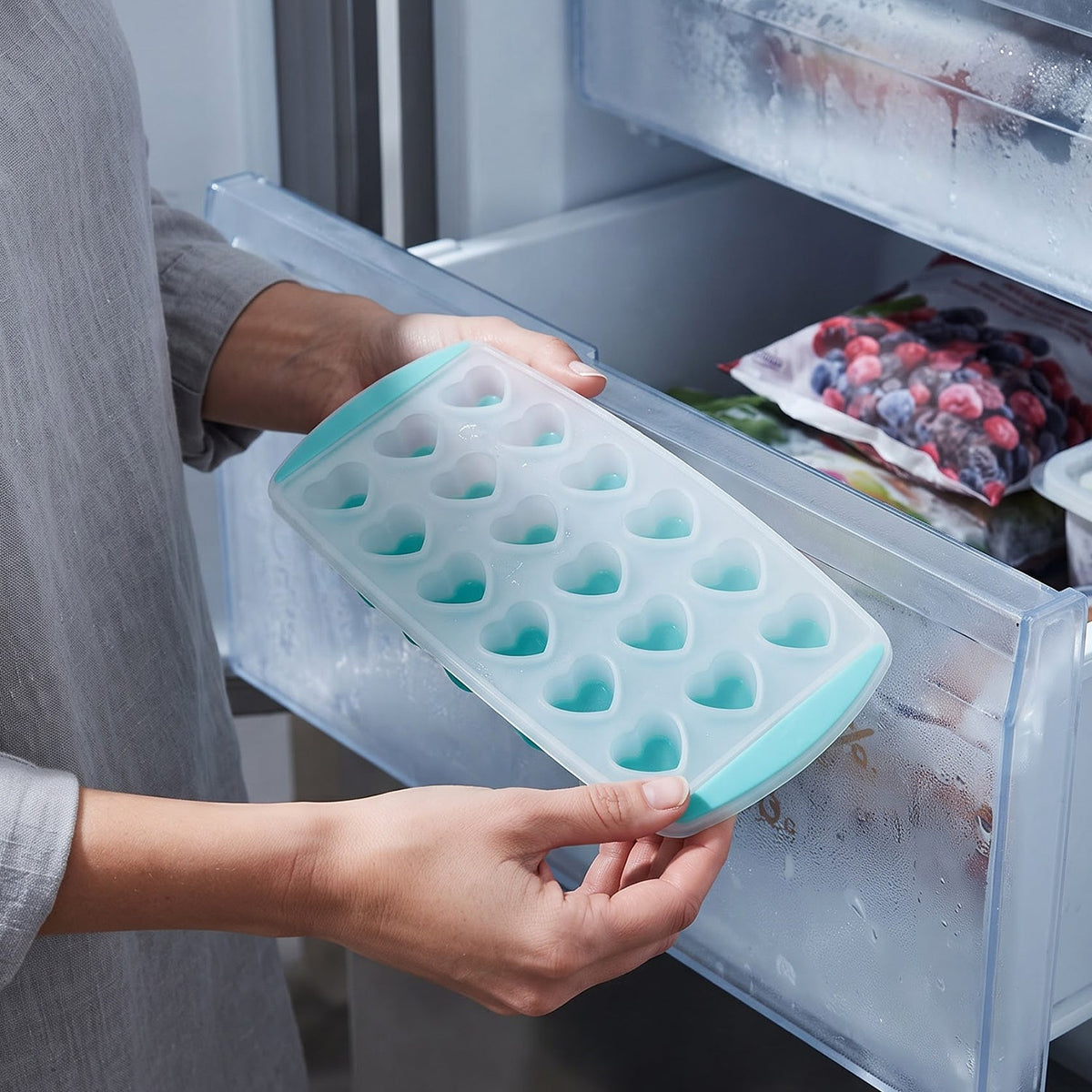 A person holds a clear and teal heart-shaped ice cube tray over an open freezer drawer, filled with frozen berries.