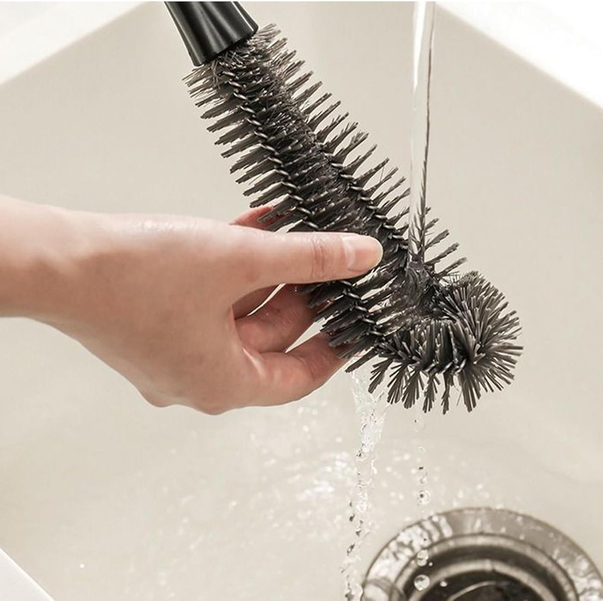 A hand holds the flexible grey brush head under running water in a white sink, showing its bristles.