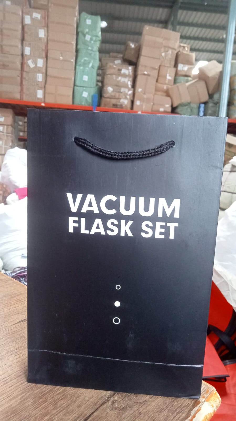 A black gift bag with 'VACUUM FLASK SET' printed on it stands on a wooden surface in a warehouse setting.