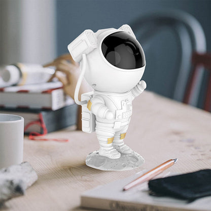 A white astronaut projector stands on a wooden desk, surrounded by books and a pen, with a white mug to the left.