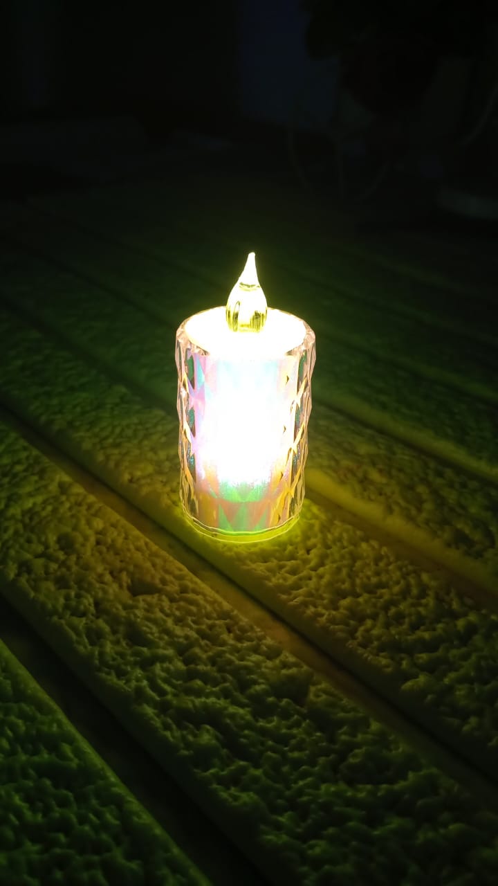 A single lit LED tea light with a textured clear body and a bright white glow, resting on a green surface.