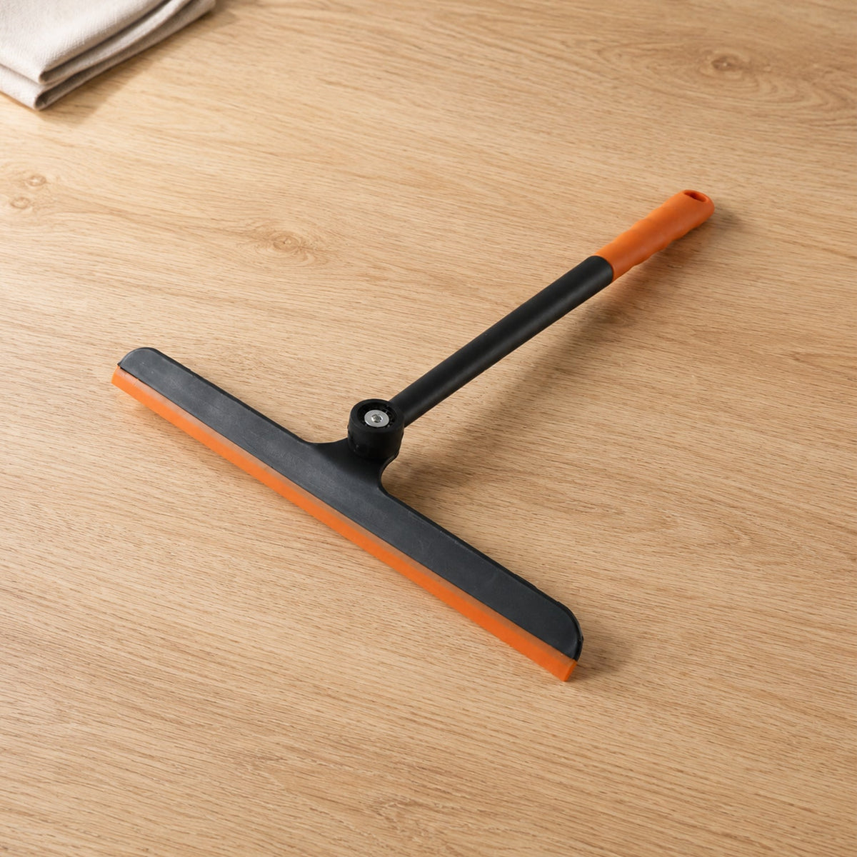 An orange and black squeegee with a pivoting head rests on a light wood surface next to a folded white towel.