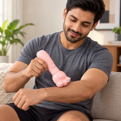 A man in a grey t-shirt uses a pink bear paw shaped massager on his upper arm while sitting on a couch.
