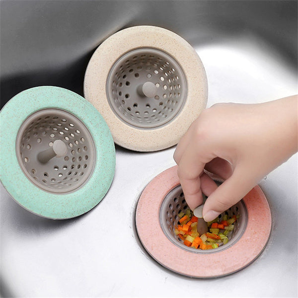 Three silicone sink strainers in mint green, beige, and pink, with a hand removing food scraps from the pink one.