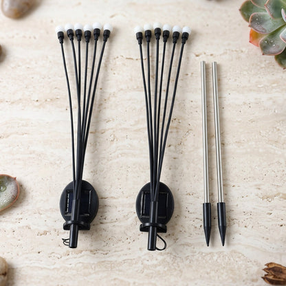 Two disassembled solar firefly lights with white bulbs and two silver stakes are arranged on a stone surface.