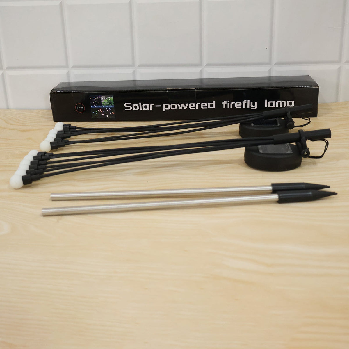Two solar firefly light units and stakes are displayed in front of a black box on a light wooden surface.