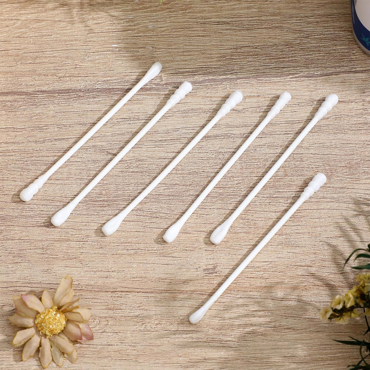 Six white cotton swabs with rounded tips are scattered on a light brown wooden surface, with a decorative flower and leaves