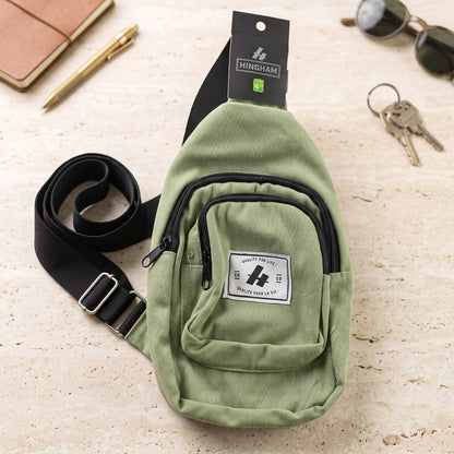 A green canvas sling bag with a black strap and zippers, lying on a light-colored textured surface with a pen, keys, and