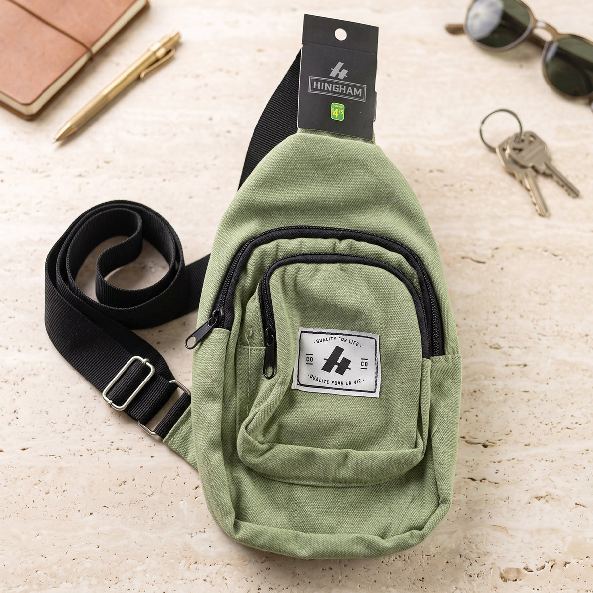 A green canvas sling bag with a black strap and front pocket lies on a textured surface next to a pen and keys.