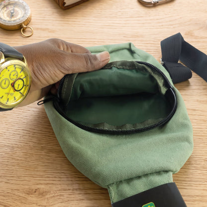 A hand opens the main zipper compartment of the green canvas sling bag, revealing its dark green lining.