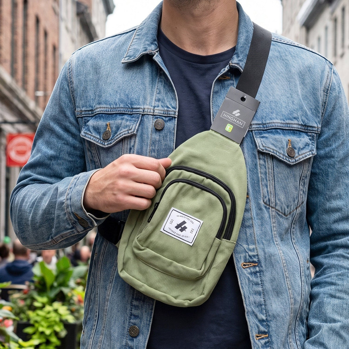 A man in a denim jacket wearing a green canvas sling bag with a black strap across his chest, on a city street.