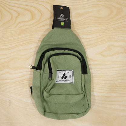 A green canvas sling bag with a black strap and zippers, featuring a white label, displayed flat on a light wood surface.