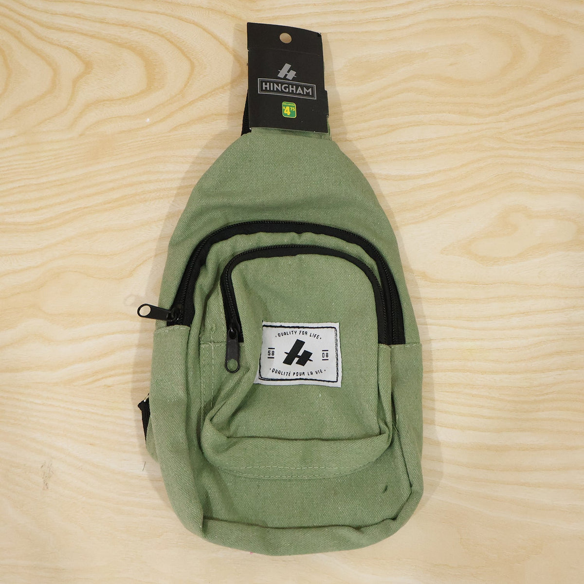 A green canvas sling bag with a black strap and front pocket is displayed on a light wooden surface.
