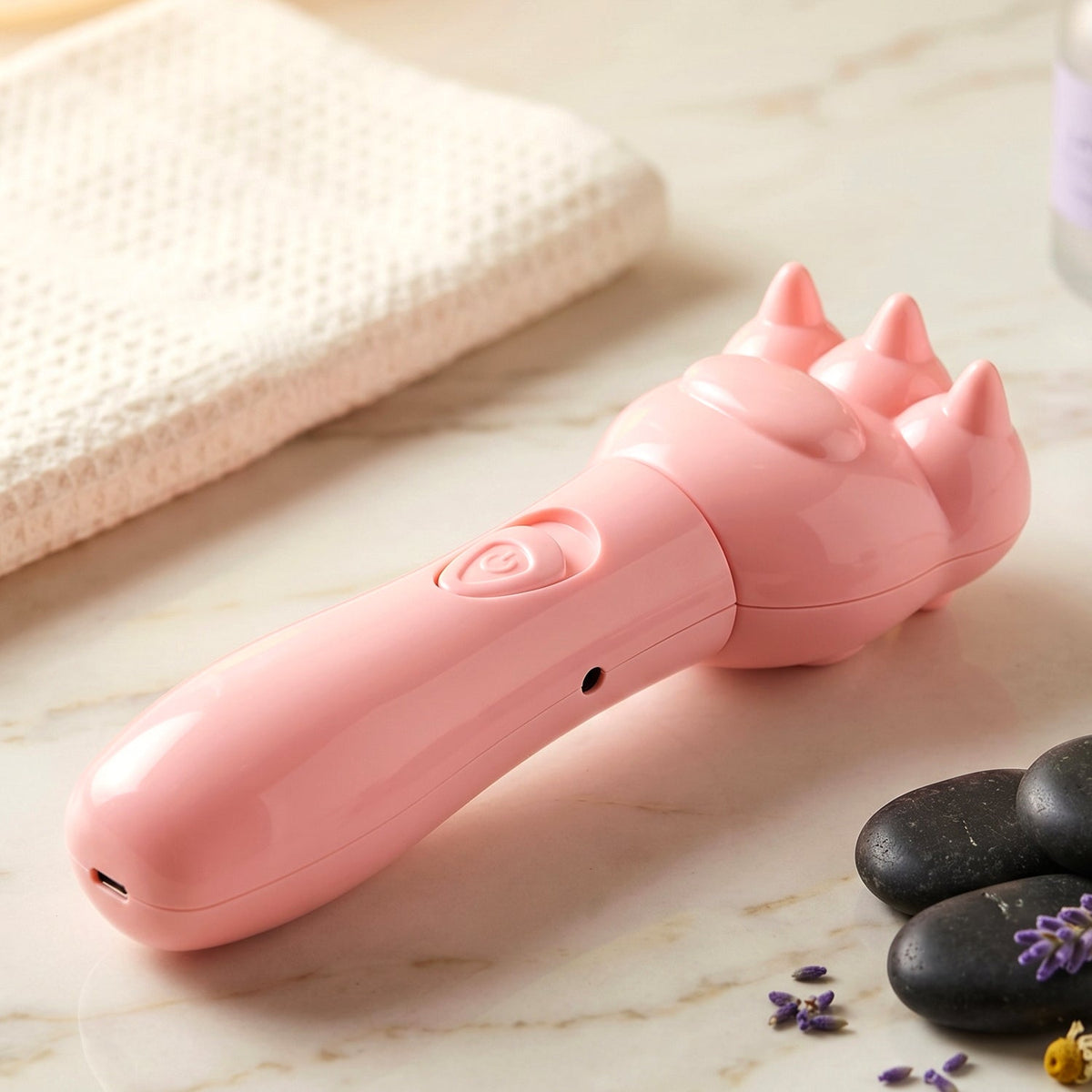 A pink bear paw massager sits on a light marble surface next to a white towel, dark stones, and lavender sprigs.