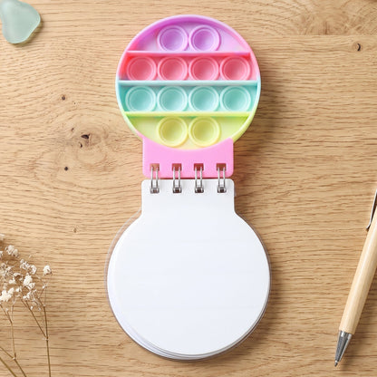 A top-down view of the rainbow pop-it fidget notebook on a wooden desk with a pen and dried flowers.