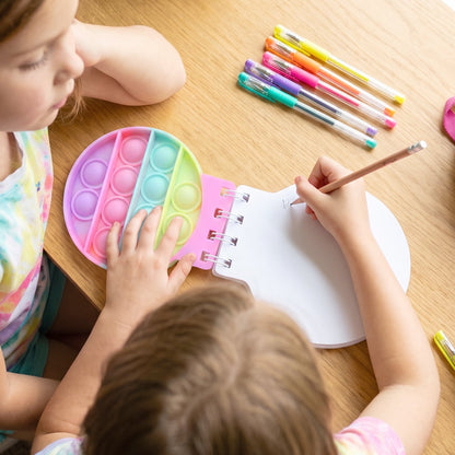Two children at a wooden table, one writing in a pop-it fidget notebook, the other pressing its bubbles.