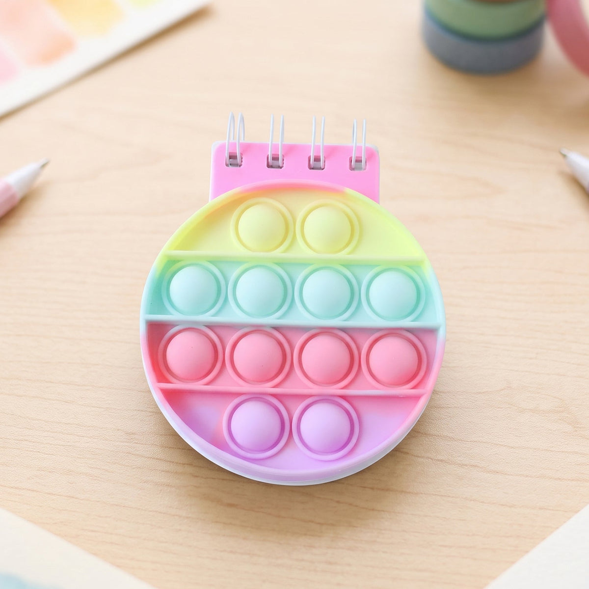A rainbow pop-it fidget notebook with a pink spiral binding and pastel colored bubbles on a light wooden desk.