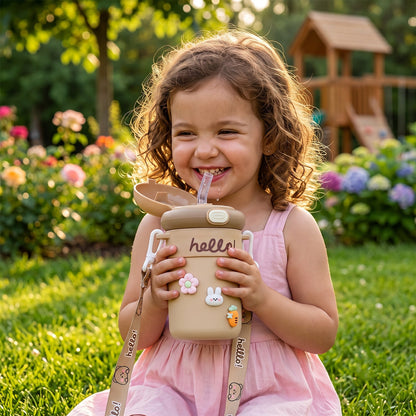 A happy child holds a tan sipper bottle with a straw, charms, and a patterned strap in a sunny garden.