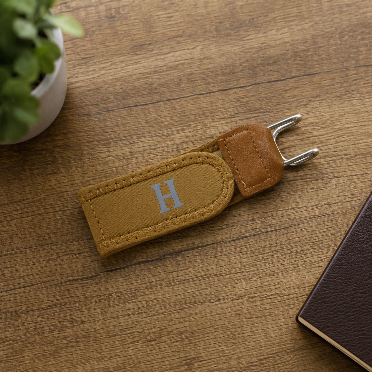 A tan buckle-less belt with a silver 'H' rests on a wooden table next to a green plant and a brown notebook.