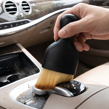 A hand holding a black brush with light brown bristles, cleaning crumbs from a car's center console.