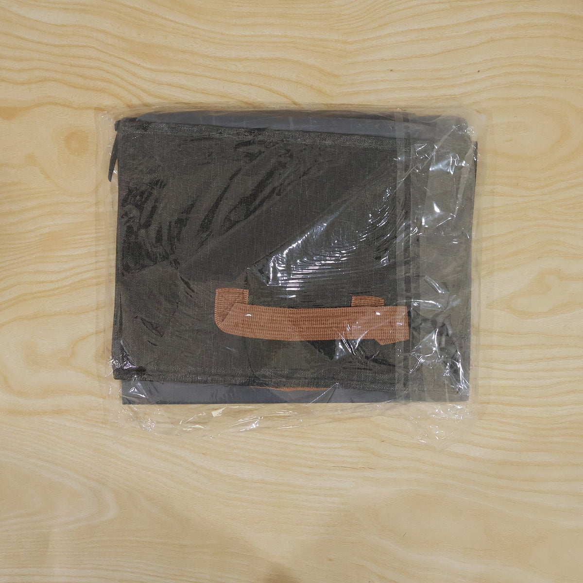 A dark gray foldable fabric storage basket with a brown handle, folded and sealed in clear plastic packaging.