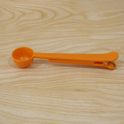 A simple orange plastic measuring spoon with a clip is positioned on a light brown wooden surface.