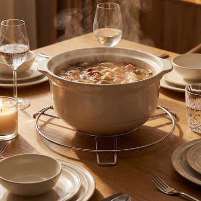 A steaming beige hot pot sits on a stainless steel stand on a wooden dining table set with glasses and plates.