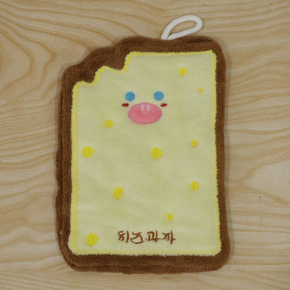 A cartoon cheese toast-shaped microfiber towel with a brown crust and yellow center, featuring a face, lies on a wooden