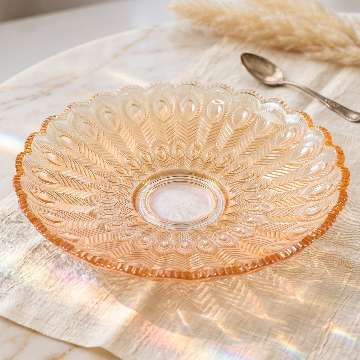 Close-up of an empty amber glass platter with an intricate feather pattern, reflecting sunlight on a white linen surface.