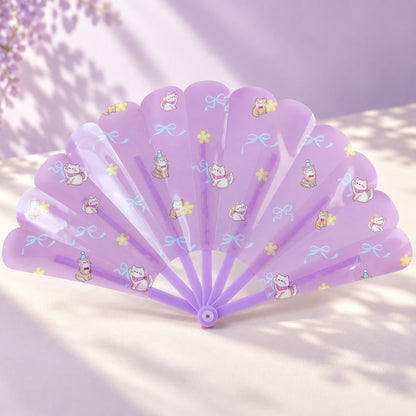 An open purple hand fan with playful cat and bow patterns rests on a light purple surface with shadows.