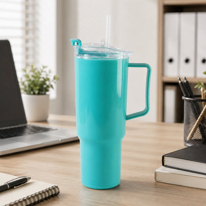 An aqua blue tumbler with a handle, clear lid, and straw sits on a wooden desk next to a laptop and office supplies.