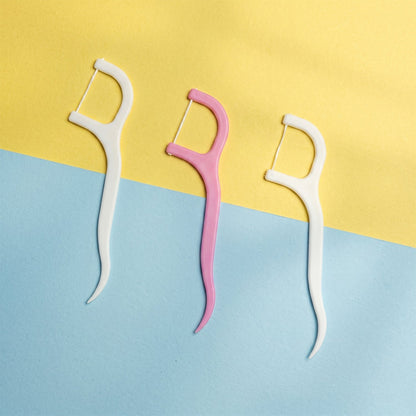 Three dental floss picks, two white and one pink, standing upright against a two-toned yellow and blue background.