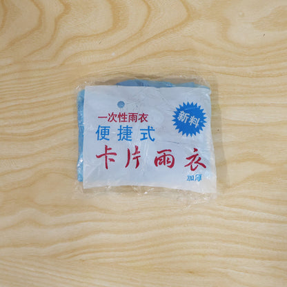 A small, clear plastic package containing a folded blue rain poncho is centered on a light wooden surface.