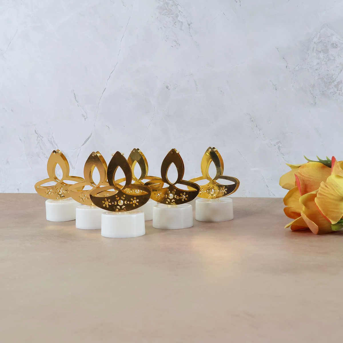Six golden LED diya tealight candles arranged on a light brown surface with a bouquet of orange flowers on the right.