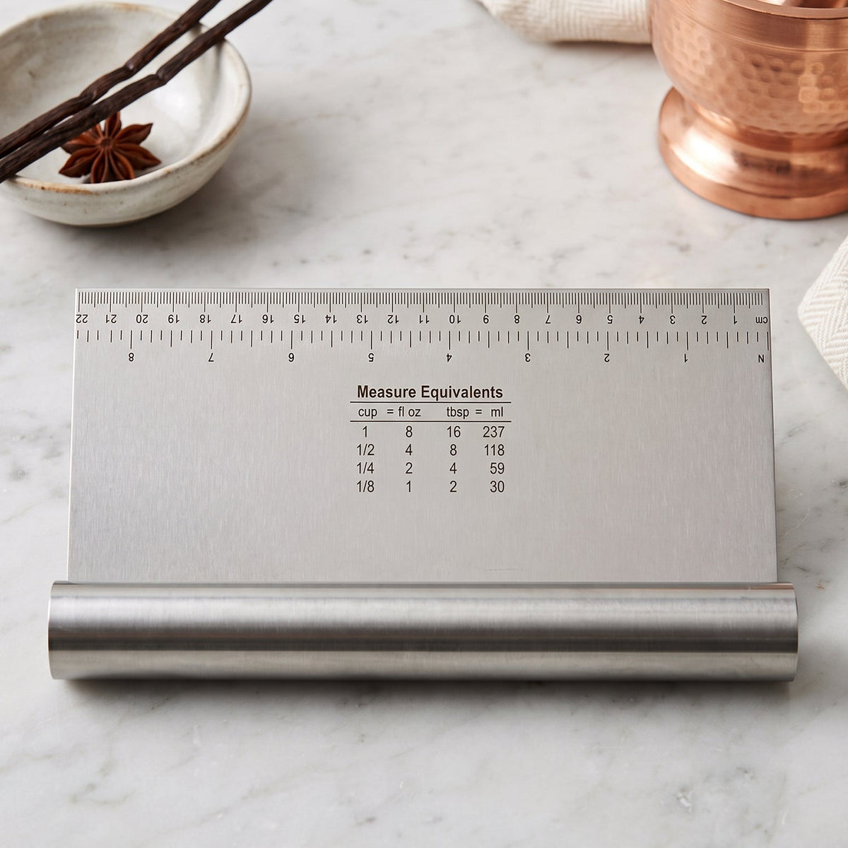 A stainless steel dough scraper with a ruler and measurement equivalents sits on a white marble surface.