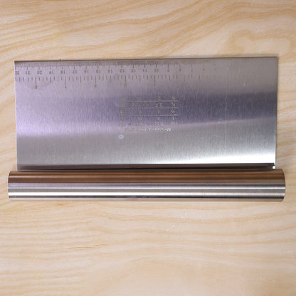 A stainless steel dough scraper with a ruler and measurement equivalents is displayed on a light wooden background.