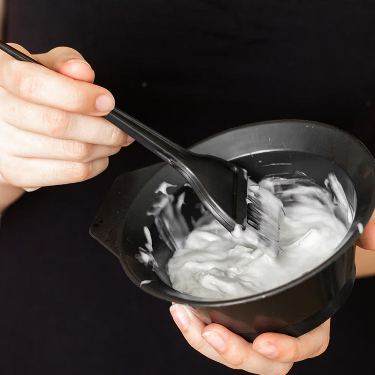 Hands holding a black bowl with white hair dye and a black brush mixing the contents.