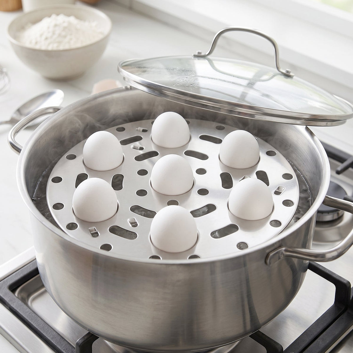 A stainless steel pot on a stove contains a steamer rack with six white eggs, steam rising, and the lid resting beside it.