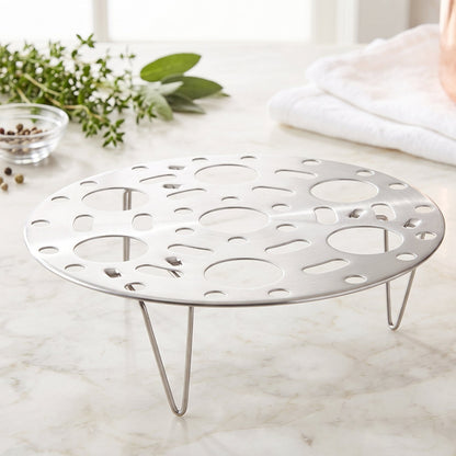 A polished metal egg steamer rack with three legs sits on a bright marble countertop, surrounded by green herbs.