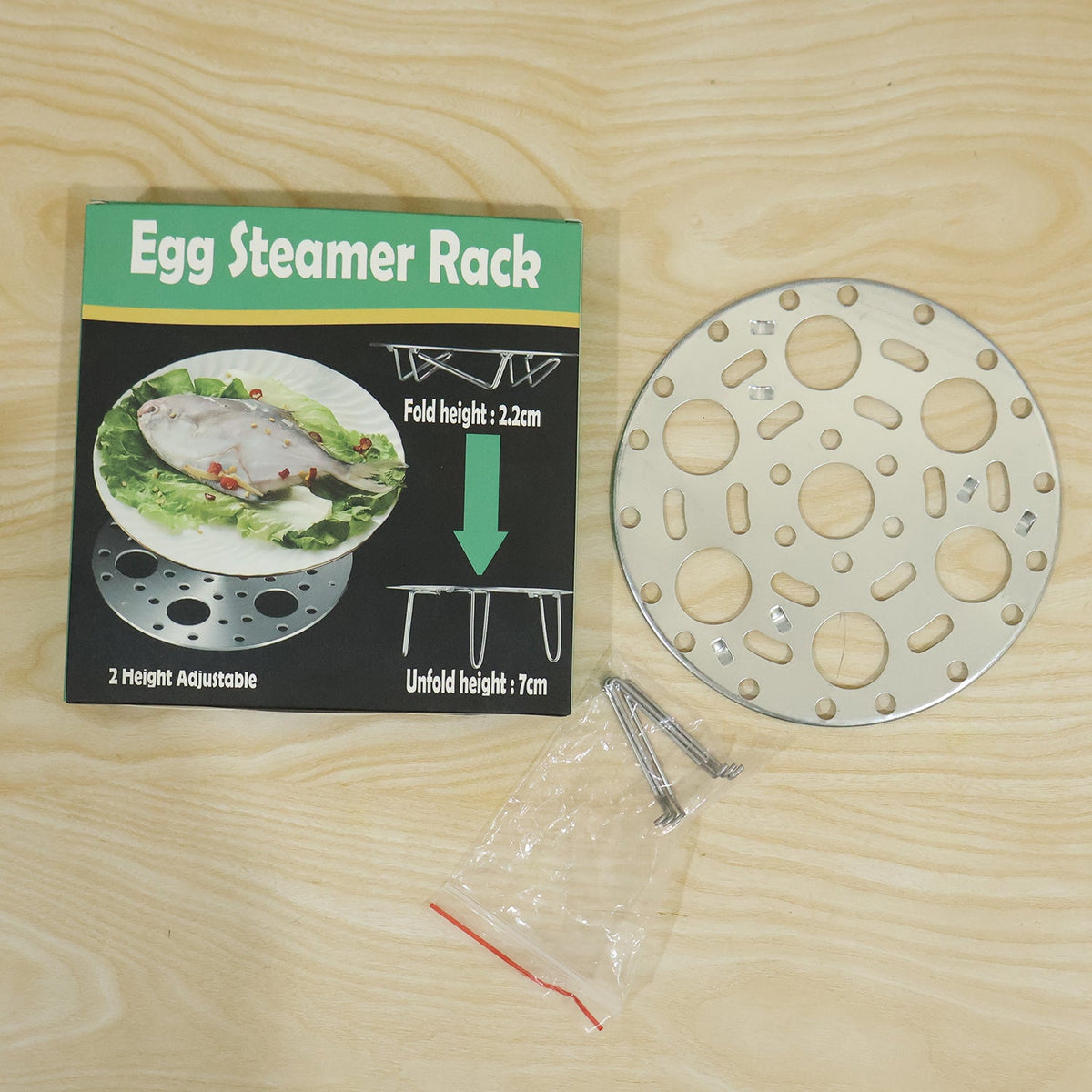 An egg steamer rack and its components, including a small bag of legs, are laid out next to its product box on a wooden