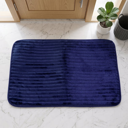 A navy blue ribbed memory foam bath mat lies on a white marble floor in front of a light wood door.